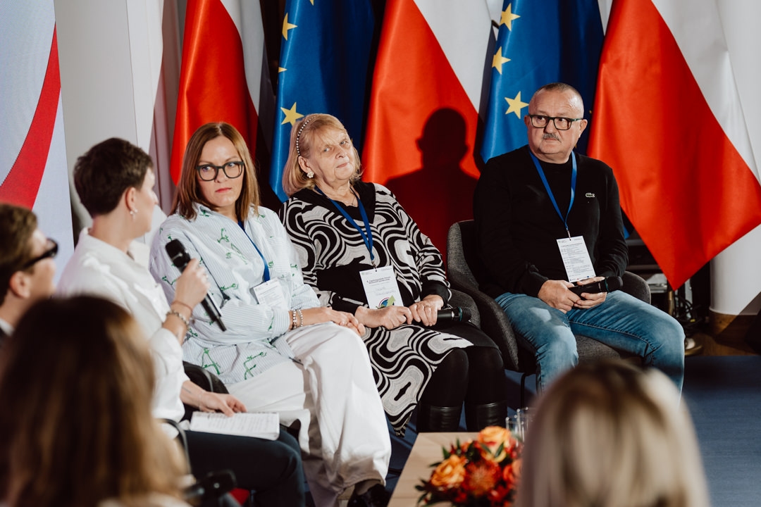 Vier Diskussionsteilnehmer sitzen mit Mikrofonen und Namensschildern auf der Bühne. Hinter ihnen sind die Flaggen von Polen und der Europäischen Union zu sehen. Sie scheinen eine Diskussion auf einer Veranstaltung oder Konferenz zu führen.  