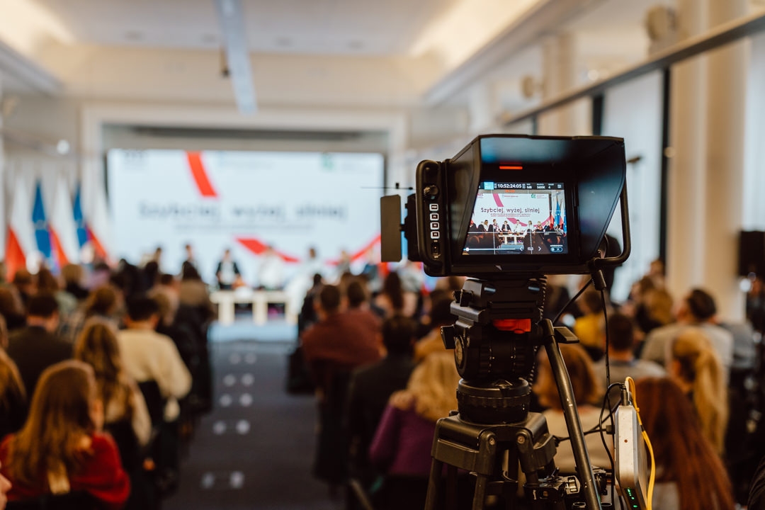 Eine Videokamera nimmt eine Podiumsdiskussion in einem Konferenzraum mit sitzenden Teilnehmern auf. Auf dem Display der Kamera sind die Redner und das Publikum zu sehen, während im Hintergrund Fahnen und der Präsentationsbildschirm zu sehen sind. 
