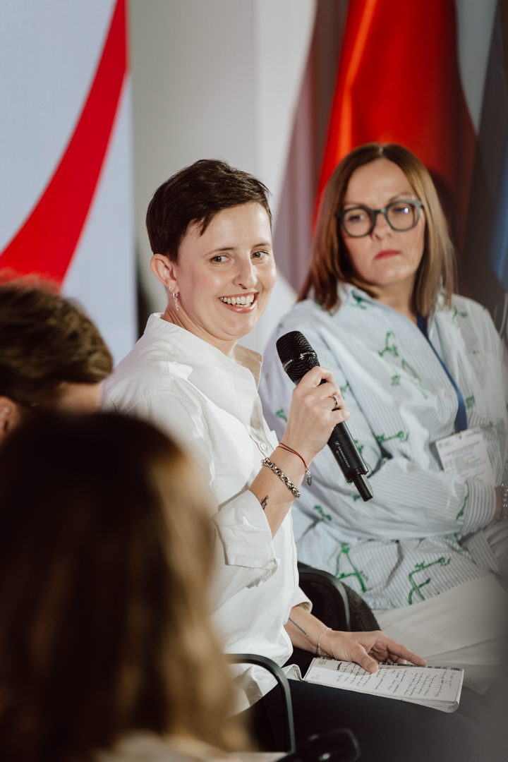 Eine Frau in einem weißen Hemd lächelt, während sie ein Mikrofon hält und während einer Podiumsdiskussion spricht. Eine andere Frau mit Brille schaut aufmerksam im Hintergrund zu. Unscharfe Menschen und Fahnen sind ebenfalls zu sehen.  