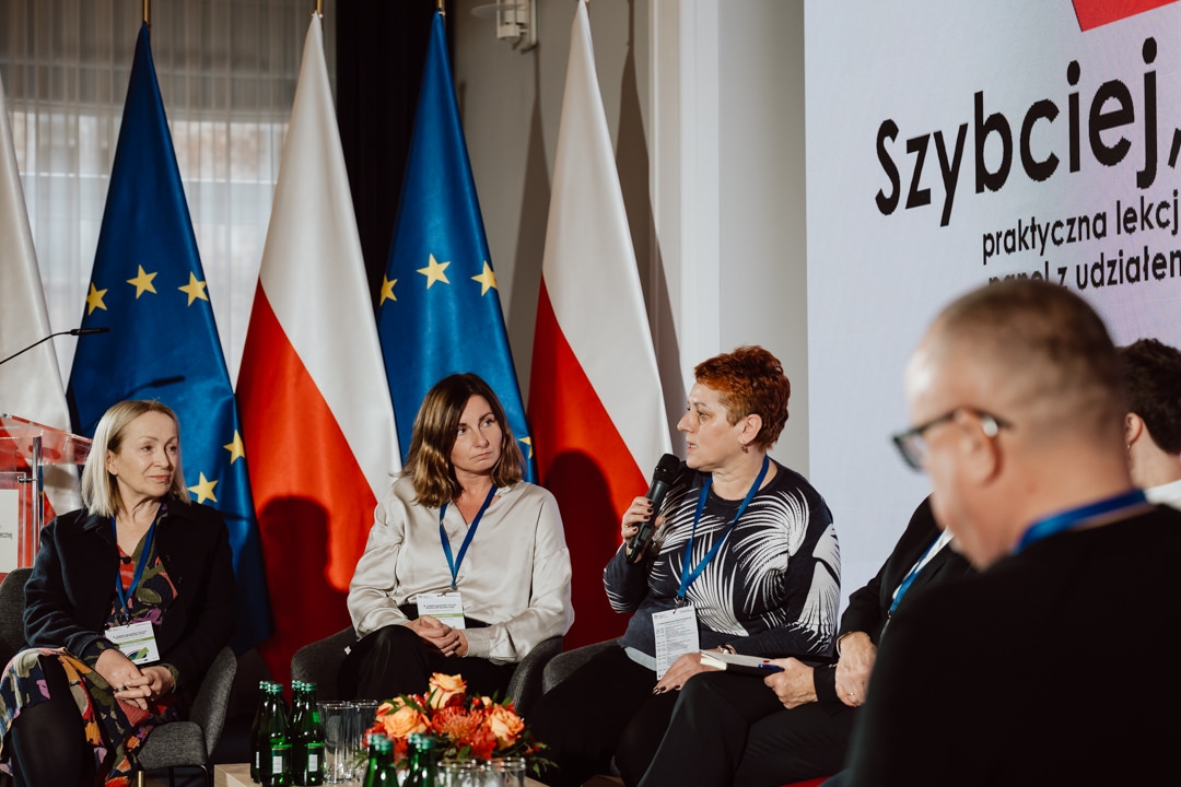 Fünf Personen sitzen während einer Podiumsdiskussion auf der Bühne, wobei drei Frauen im Rampenlicht stehen. Eine der Frauen spricht in ein Mikrofon. Hinter ihnen sind die Flaggen Polens und der Europäischen Union zu sehen und ein großer Bildschirm mit polnischem Text. Auf einem Tisch davor sind Flaschen zu sehen.   