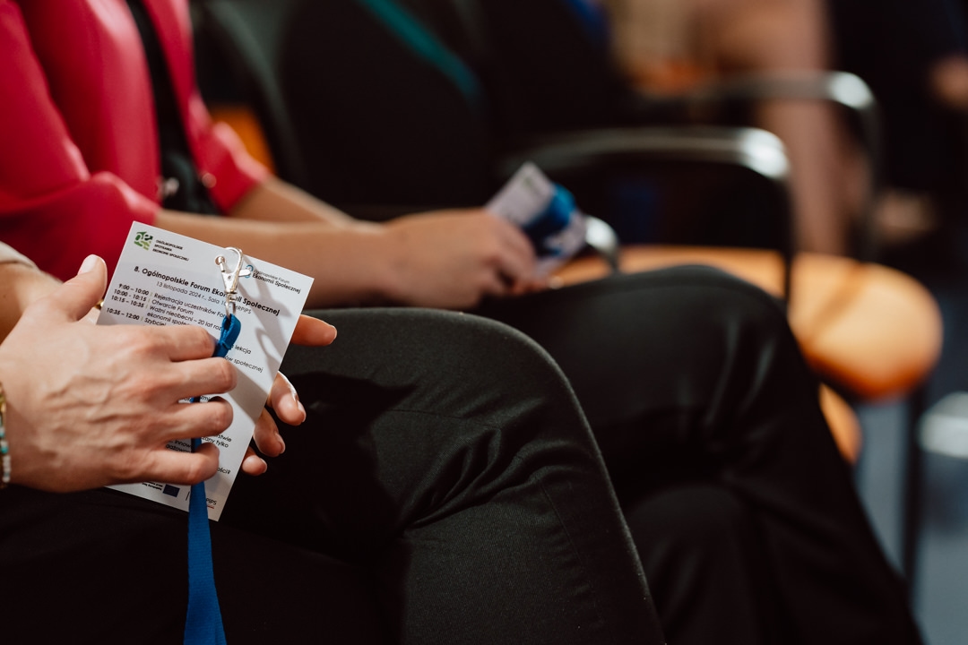 Nahaufnahme einer sitzenden Person, die ein Namensschild und ein Veranstaltungsprogramm in der Hand hält, mit einer weiteren unscharfen Person im Hintergrund, die eine Konferenz oder ein Seminar andeutet.