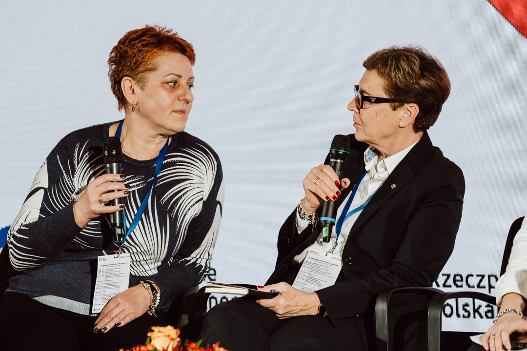 Zwei Frauen mit Konferenzschildern sitzen während einer Podiumsdiskussion und sprechen in Mikrofone. Die eine trägt einen gemusterten Pullover, die andere eine dunkle Jacke und eine Brille. Beide scheinen in ein Gespräch vertieft zu sein.  