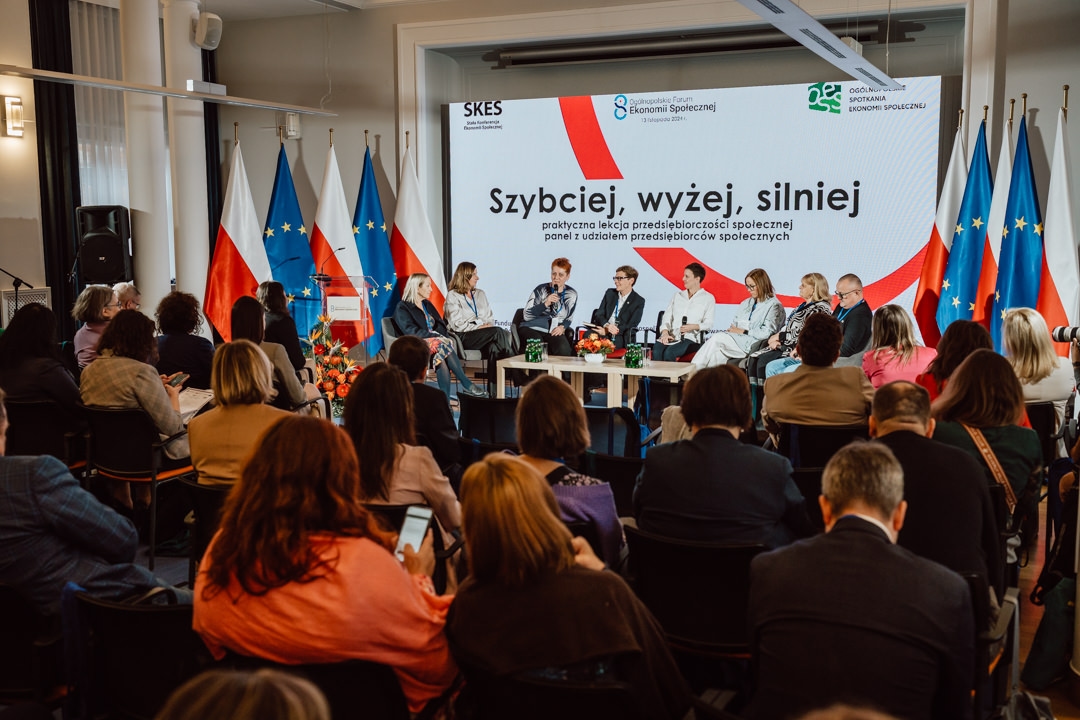 Eine Gruppe von Rednern sitzt auf einer Bühne vor dem Publikum der Konferenz. Hinter ihnen sind polnische und EU-Flaggen und ein großer Bildschirm mit polnischem Text zu sehen. Die Menschen im Publikum hören zu und machen sich Notizen.  