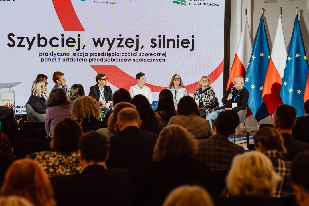 Eine Gruppe von Rednern sitzt auf einer Bühne vor dem Publikum und diskutiert über soziales Unternehmertum. Hinter ihnen ist ein großer Bildschirm mit polnischem Text und mehreren nationalen und EU-Flaggen zu sehen. 