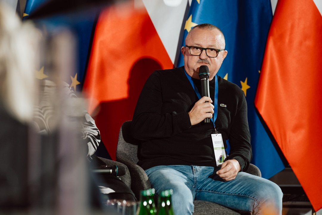 Ein Mann mit Brille und schwarzem Pullover spricht in ein Mikrofon, während er auf der Bühne sitzt. Hinter ihm sind die Flaggen der Europäischen Union und Polens zu sehen. Im Vordergrund sind Flaschen zu sehen.  