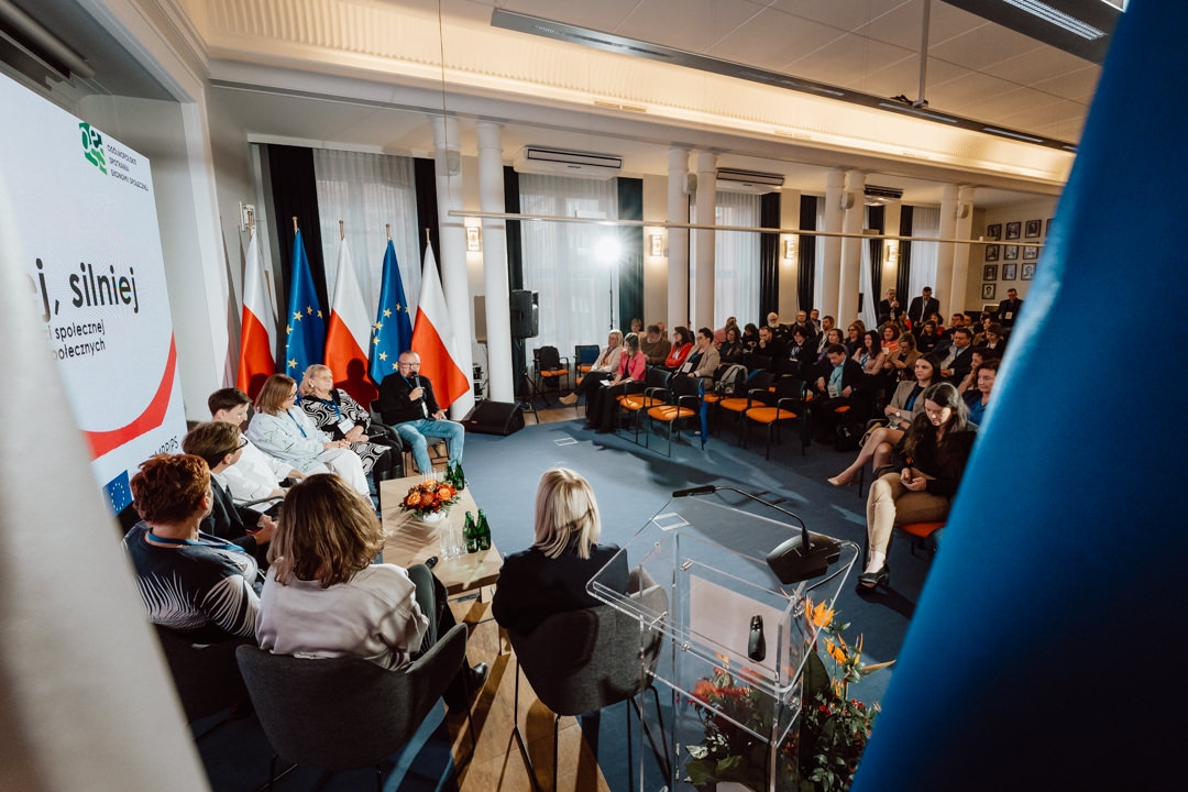In einem Konferenzraum mit polnischen und EU-Flaggen, Blumenschmuck und einer Präsentationsleinwand spricht eine Gruppe von Rednern zu den Zuhörern. Die Teilnehmer sitzen und hören aufmerksam zu. 