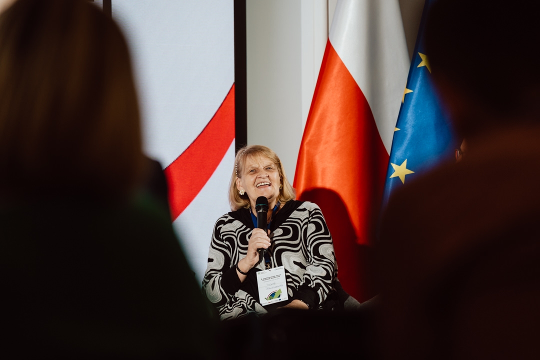 Eine Frau, die ein Mikrofon hält, lächelt, während sie auf einer Veranstaltung spricht. Sie trägt einen gemusterten Pullover und sitzt vor den Flaggen von Polen und der Europäischen Union. Im Vordergrund sind Silhouetten des Publikums zu sehen.  