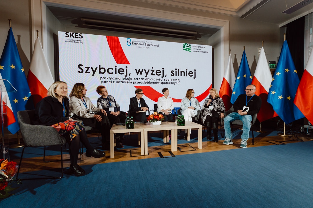Sieben Personen sitzen während einer Podiumsdiskussion auf einer Bühne vor einem großen Bildschirm, auf dem polnische Texte angezeigt werden. Im Hintergrund sind polnische und EU-Flaggen zu sehen. Die Atmosphäre wirkt formell und professionell.  