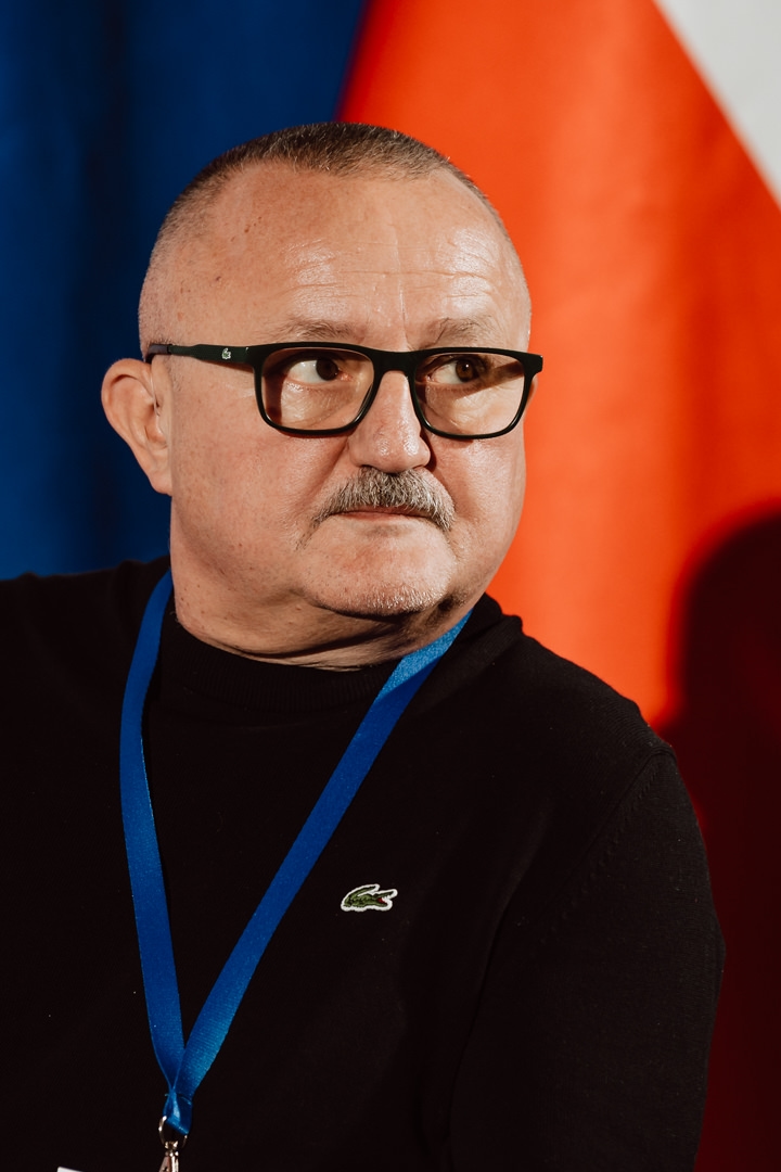 Ein Mann mittleren Alters mit Brille und Schnurrbart, der einen schwarzen Pullover mit einem blauen Schlüsselband trägt, sitzt vor einem verschwommenen Hintergrund aus Rot und Blau.