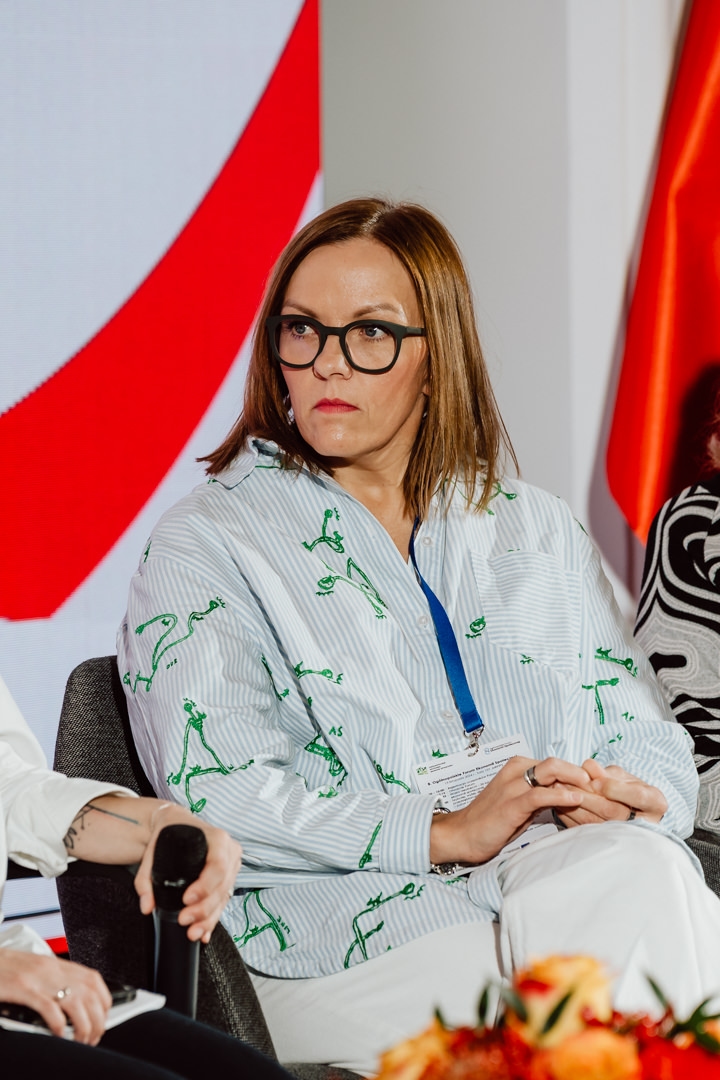 Eine Frau mit schulterlangem hellbraunem Haar und Brille sitzt auf einem Stuhl und hört aufmerksam zu. Sie trägt ein gestreiftes Hemd mit grünen Mustern, eine weiße Hose und ein blaues Schlüsselband. Hinter ihr ist ein farbenfroher Hintergrund zu sehen.  