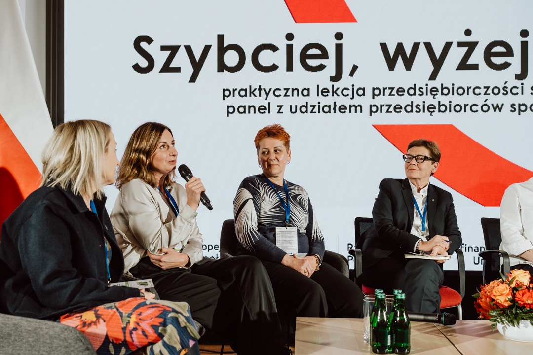 Vier Frauen sitzen und nehmen an einer Podiumsdiskussion teil, eine spricht in ein Mikrofon. Hinter ihnen zeigt ein Bildschirm einen polnischen Text an. Auf dem Tisch vor ihnen liegen drei grüne Wasserflaschen und Papiere.  