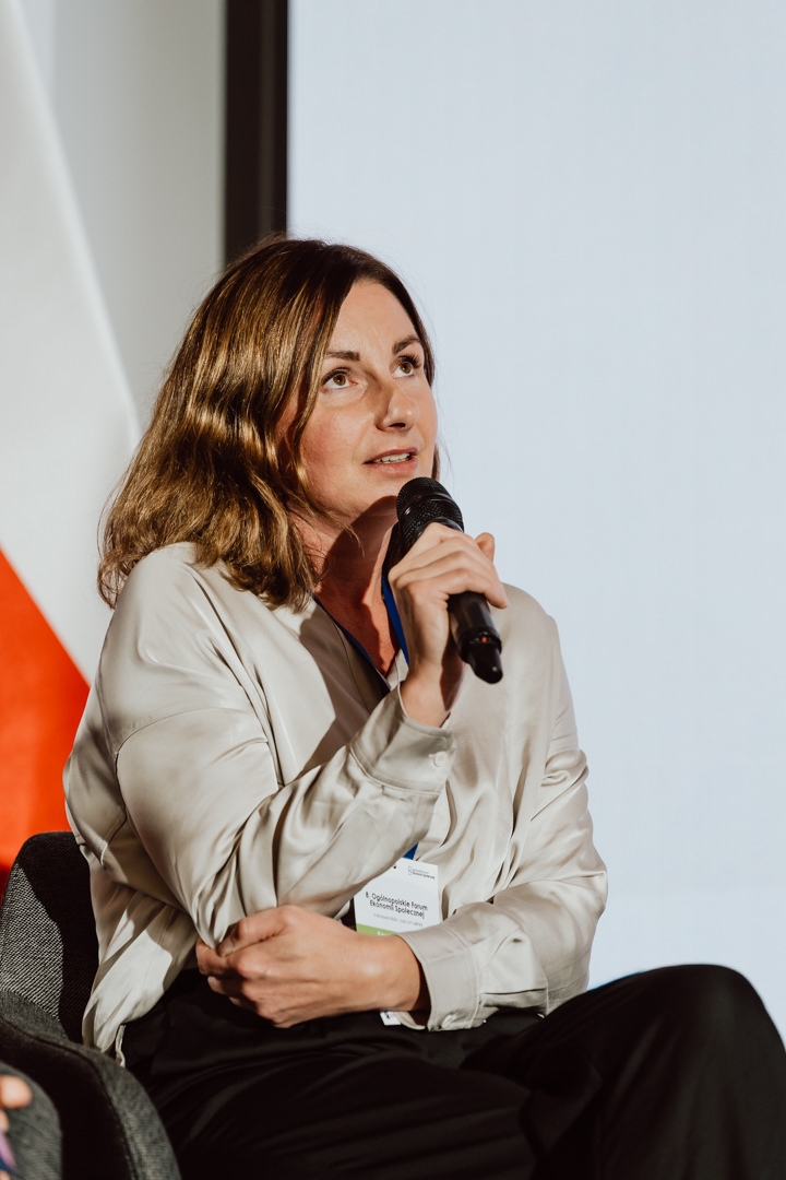 Eine Frau mit schulterlangen braunen Haaren, die eine helle Bluse trägt und ein Mikrofon in der Hand hält, sitzt auf einer Veranstaltung oder Konferenz und spricht. Ein Namensschild ist auf ihrer Bluse zu sehen. 