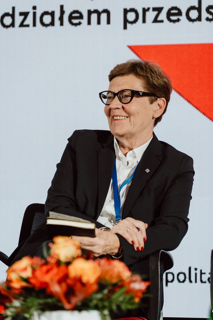 Eine Frau im Anzug und mit Brille sitzt lächelnd da und hält einen Notizblock. Im Vordergrund ist ein Strauß orangefarbener Blumen zu sehen und hinter ihr steht ein großer Text auf weißem Hintergrund. 