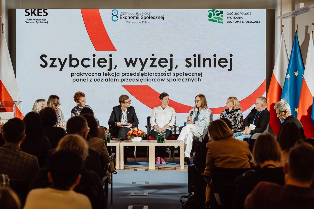 Eine Gruppe von acht Personen sitzt mit Mikrofonen auf der Bühne und diskutiert vor einem großen Bildschirm, auf dem der polnische Text zum Forum der Sozialwirtschaft angezeigt wird. Das Publikum schaut zu und auf der rechten Seite sind Fahnen zu sehen. 