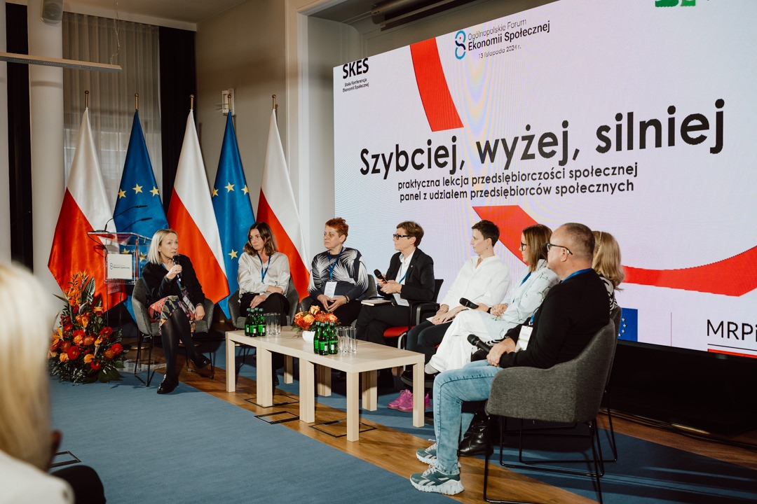 Eine Gruppe von sechs Personen sitzt auf einer Bühne und diskutiert vor einem Publikum. Hinter ihnen sind die Flaggen Polens und der EU zu sehen, und auf einem Bildschirm wird ein Text in polnischer Sprache angezeigt. Auf einem Tisch vor ihnen stehen Wasserflaschen und Mikrofone.  