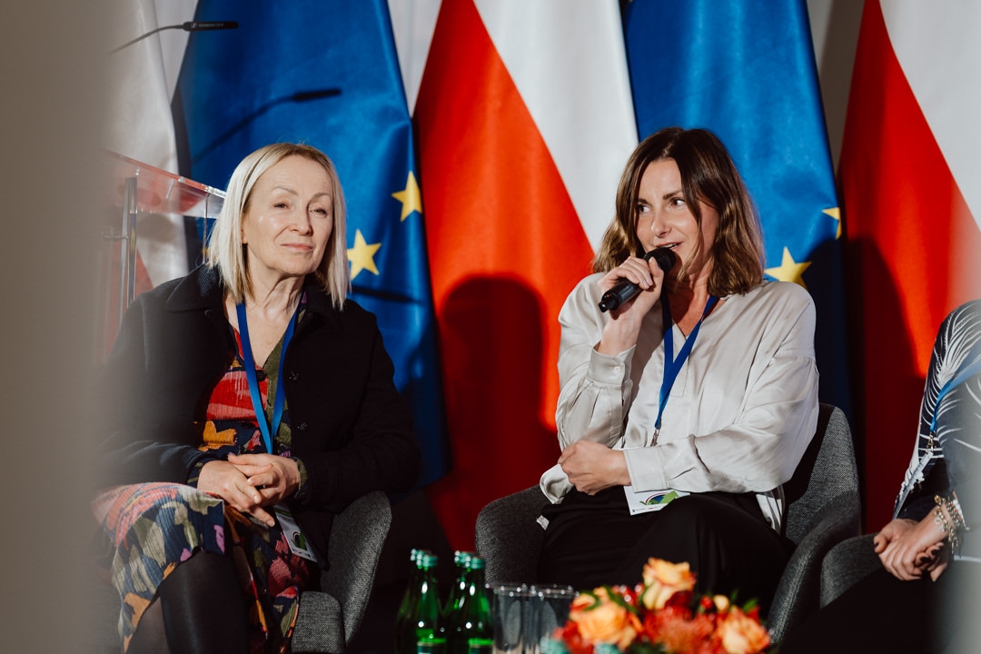 Zwei Frauen sitzen und sprechen während einer Podiumsdiskussion, eine hält ein Mikrofon. Sie tragen Konferenz-Lanyards und sitzen vor EU- und polnischen Flaggen. Auf einem Tisch im Vordergrund liegen Glasflaschen und Blumen.  