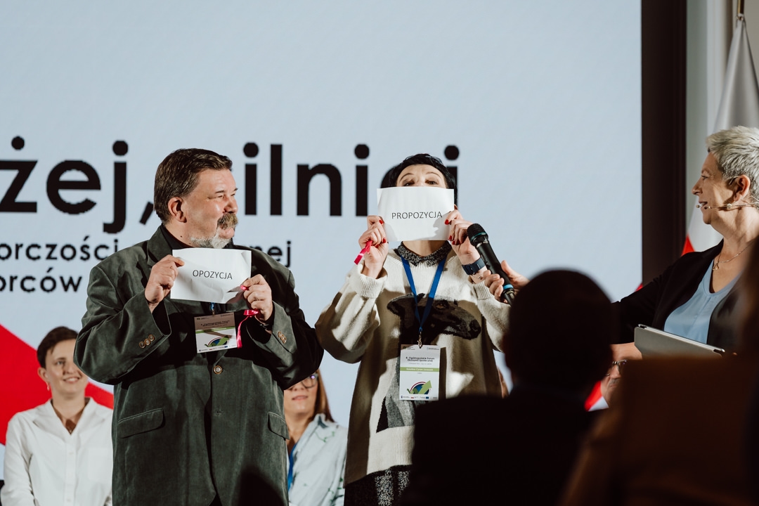 Zwei Personen auf der Bühne halten Karten mit polnischen Wörtern; eine Frau auf der rechten Seite hält ein Mikrofon und lächelt. Im Hintergrund sehen Sie mehrere Personen sitzen und einen großen Bildschirm mit Textausschnitten. 