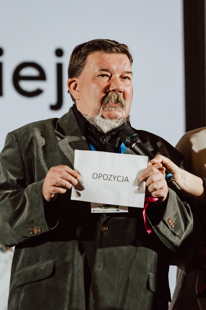 Ein Mann in einer grauen Jacke hält ein Schild mit der Aufschrift 'OPPOSITION', während er bei einer Indoor-Veranstaltung in ein Mikrofon spricht.