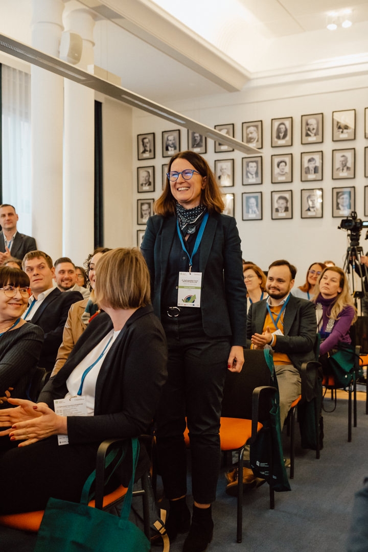 Eine Frau steht und lächelt in einem Raum voller sitzender Menschen, die applaudieren. Sie trägt eine Brille, ein Halstuch und ein Namensschild. Gerahmte Porträts bedecken die Wand hinter ihr und die Atmosphäre wirkt heiter und professionell.  