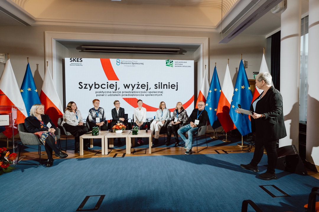 Eine Gruppe von sieben Personen sitzt auf einer Bühne mit einem Moderator, der ein Mikrofon hält. Hinter ihnen befindet sich ein Bildschirm mit polnischem Text und den Nationalflaggen Polens und der EU auf beiden Seiten. Der Raum ist hell erleuchtet und modern dekoriert.  