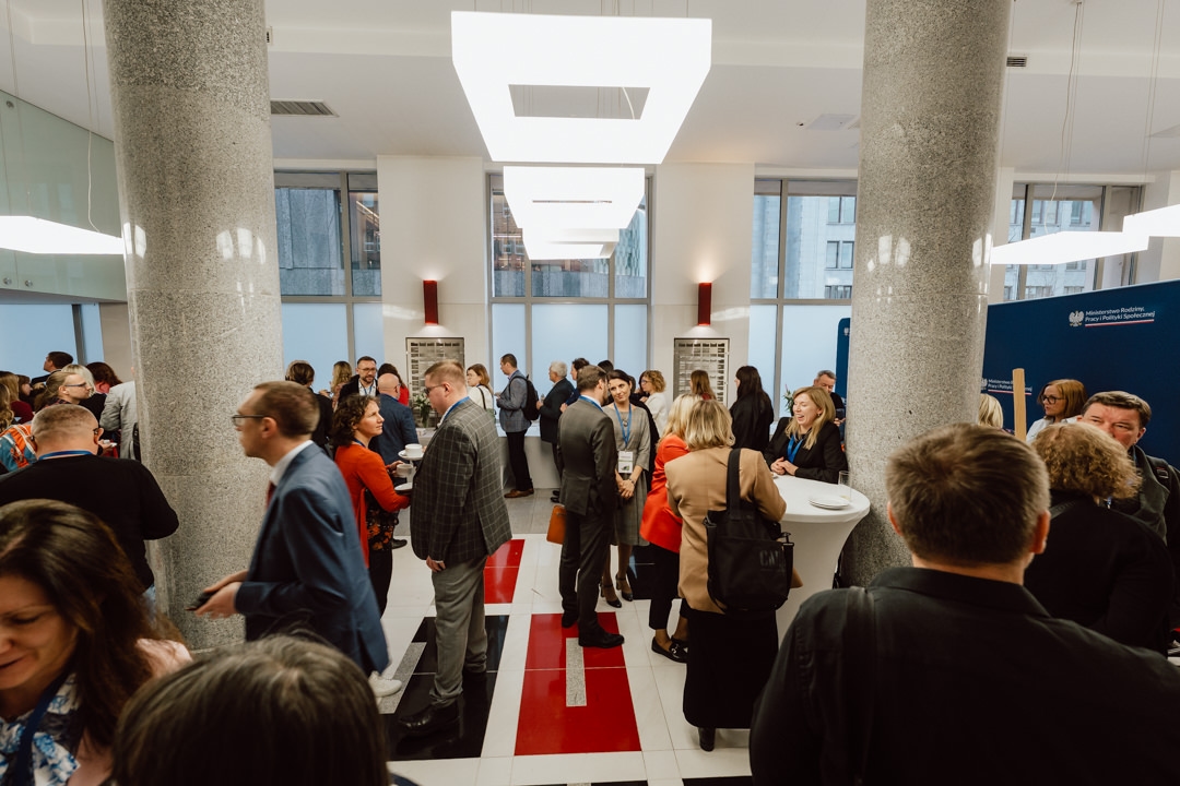 Eine Gruppe von Menschen in Geschäftskleidung, die sich bei einer Veranstaltung in einer Halle mit hohen Säulen, moderner Beleuchtung und großen Fenstern im Hintergrund treffen und Kontakte knüpfen. Einige stehen an Stehtischen, halten Getränke in der Hand und unterhalten sich. 