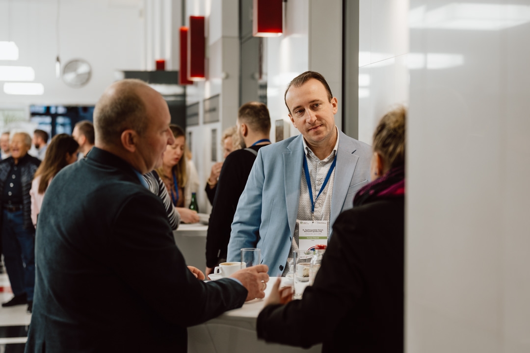 Ein Mann in einer hellblauen Jacke mit Namensschild unterhält sich während eines beruflichen Networking-Treffens mit zwei Personen an einem Stehtisch; andere Teilnehmer sind im Hintergrund zu sehen.
