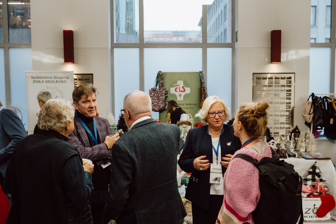 Eine Gruppe von Menschen in Geschäftskleidung plaudert und knüpft Kontakte bei der Veranstaltung in einem Raum mit Ausstellungstischen mit handgefertigtem Kunsthandwerk und Informationsmaterial im Hintergrund.