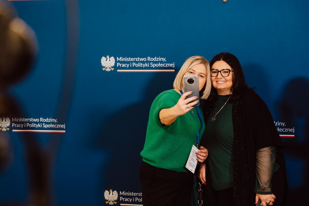 Zwei Frauen lächeln und posieren für ein Selfie vor einem blauen Hintergrund mit dem Logo und Namen des polnischen Ministeriums für Familie, Arbeit und Sozialpolitik.