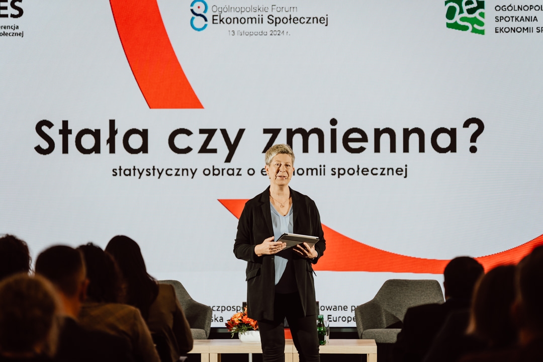 Eine Person mit einem Tablet steht auf der Bühne und spricht vor einem großen Bildschirm, auf dem der polnische Text "Fix oder variabel?" angezeigt wird, mit dem Logo des Nationalen Forums für Sozialwirtschaft im Hintergrund.