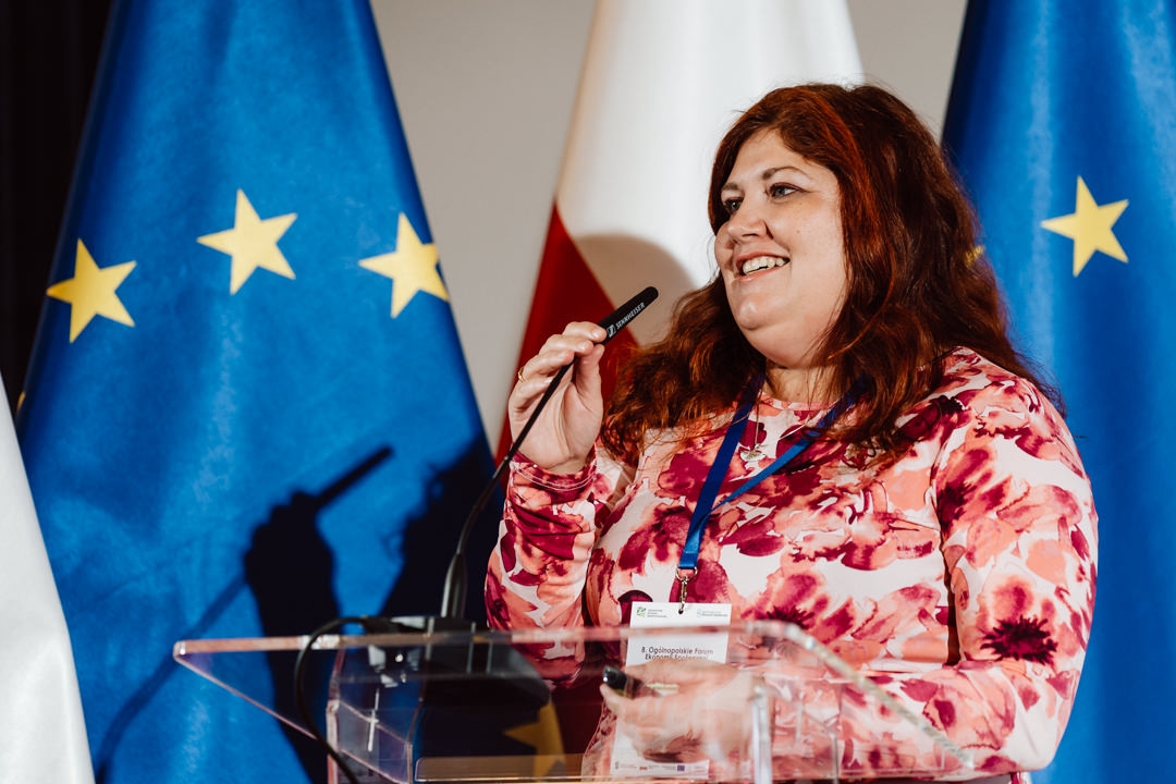 Eine Frau mit langen braunen Haaren, die ein rosa geblümtes Oberteil und ein Schlüsselband trägt, lächelt, während sie in ein Mikrofon auf einem transparenten Podium spricht. Hinter ihr wehen blaue Fahnen mit gelben Sternen und eine rot-weiße Flagge. 