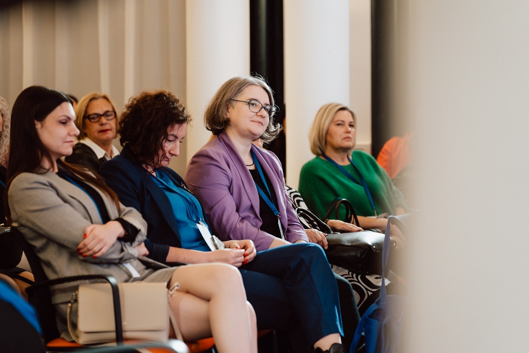Mehrere Frauen sitzen in Stuhlreihen bei einer Indoor-Veranstaltung und hören aufmerksam zu. Die meisten sind in Geschäftskleidung gekleidet und sehen konzentriert aus, was auf eine professionelle Umgebung oder eine Konferenz schließen lässt. 