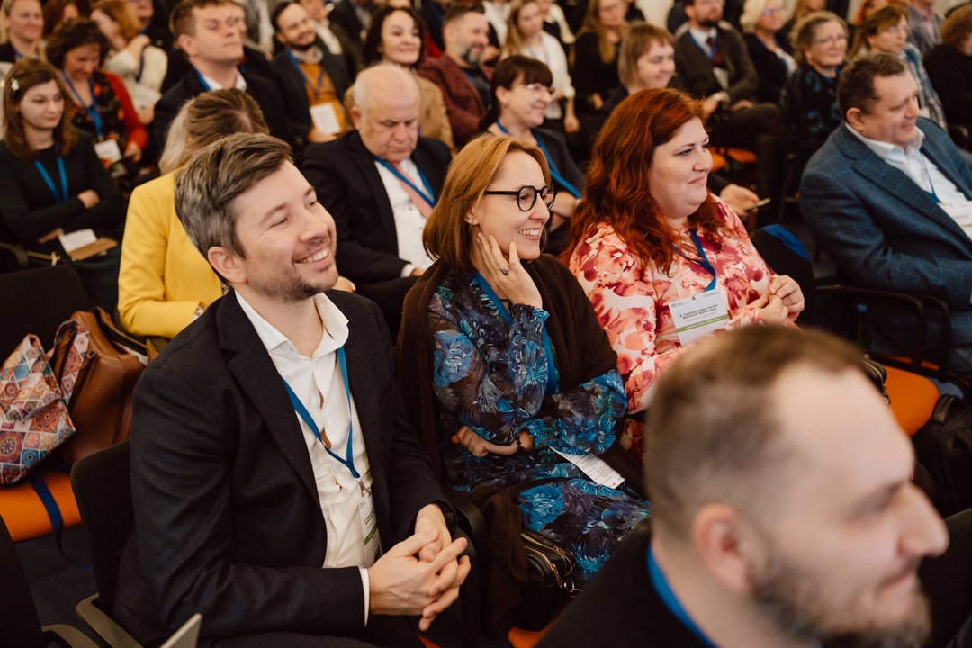 Eine Gruppe von Menschen, sowohl Männer als auch Frauen, sitzen dicht beieinander in Reihen, lächeln und hören aufmerksam zu, was eine Konferenz oder ein Seminar zu sein scheint. Die meisten von ihnen tragen Anstecker und Schlüsselbänder. 