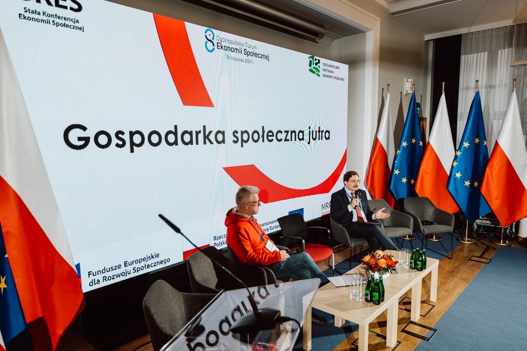 Zwei Männer sitzen auf einer Bühne mit Mikrofonen und führen eine Diskussion. Hinter ihnen ist ein großer Bildschirm mit polnischen Texten und Logos zu sehen. Vier polnische Flaggen und drei EU-Flaggen stehen rechts von der Bühne.  