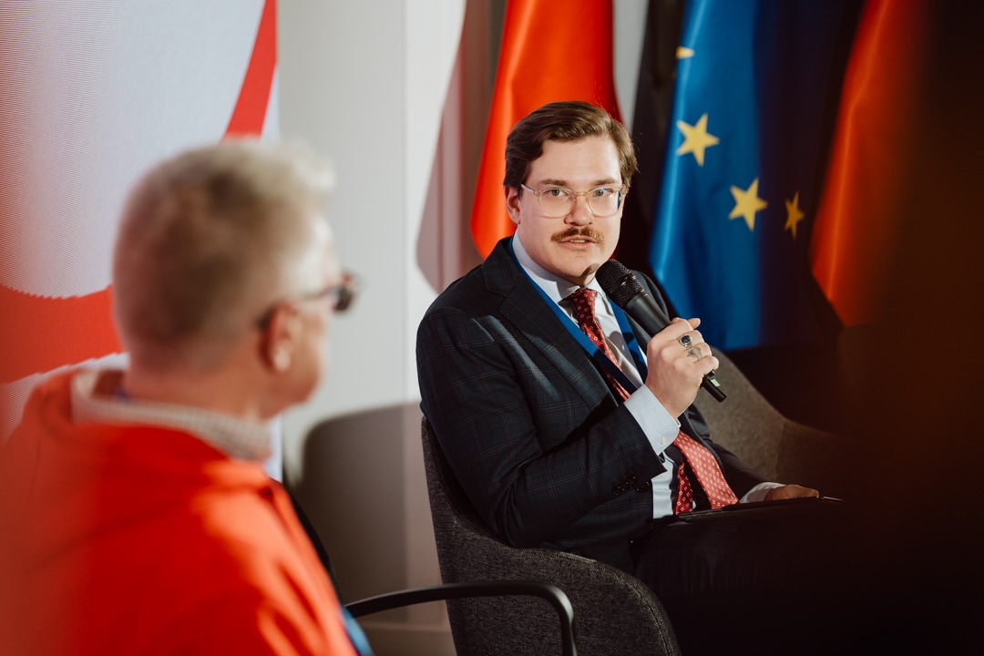 Ein Mann in einem Anzug hält ein Mikrofon und spricht während einer Podiumsdiskussion, während eine andere Person in einer roten Jacke zuhört. Im Hintergrund sind rote Flaggen der Europäischen Union zu sehen. 
