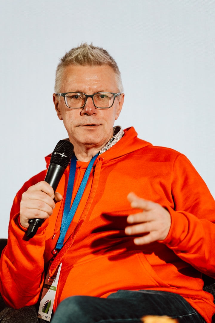 Ein Mann mit kurzen grauen Haaren und Brille, der ein leuchtend orangefarbenes Kapuzensweatshirt und ein blaues Schlüsselband trägt, spricht in ein Mikrofon, während er in einem schlichten Hintergrund sitzt.