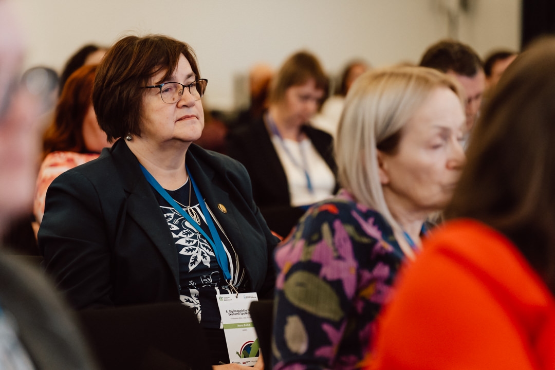 Eine Gruppe von Menschen sitzt aufmerksam in einem Konferenzraum. Im Mittelpunkt steht eine Frau mit Brille und dunkler Jacke, die einen Ausweis und ein Schlüsselband trägt und einer Präsentation zuhört. Andere sitzen in der Nähe, einige verschwommen im Vordergrund.  