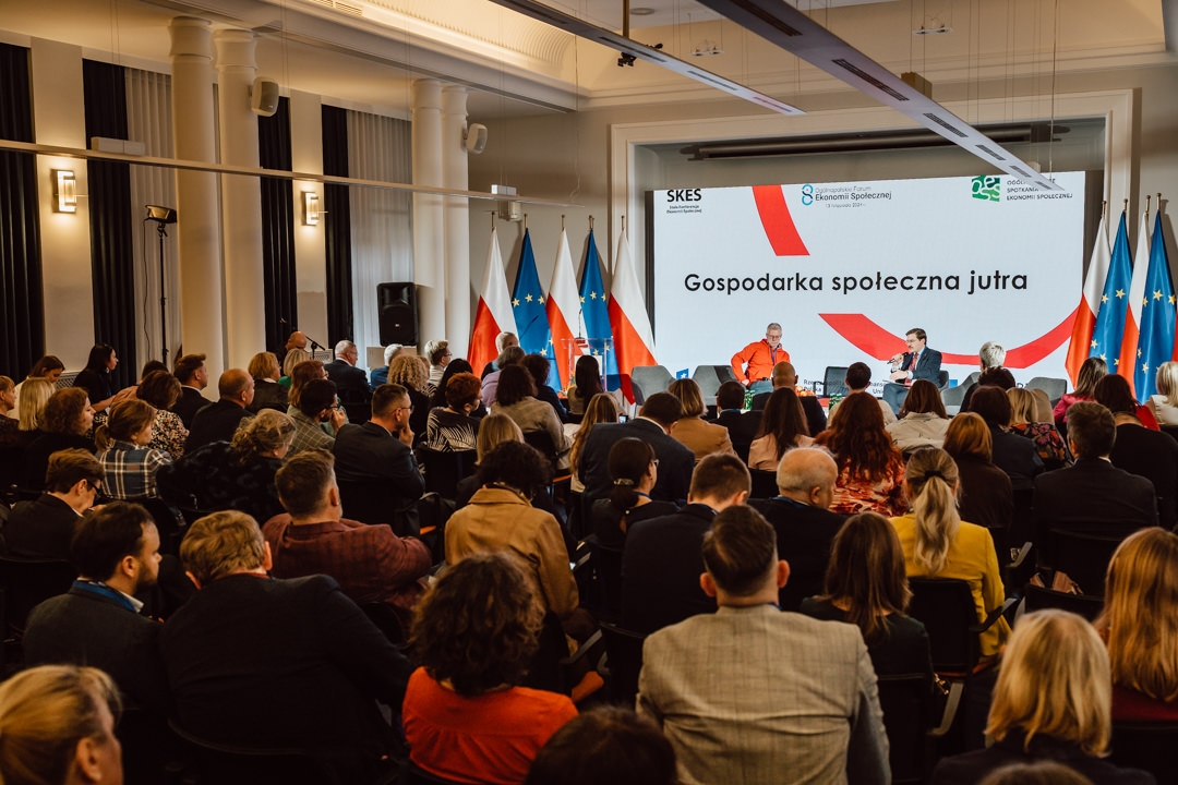 Ein großes Publikum sitzt gegenüber der Bühne, auf der die Redner an einer Podiumsdiskussion teilnehmen. Hinter ihnen wird der polnische Text 'Die Sozialwirtschaft von morgen' auf eine Leinwand projiziert. Die Flaggen von Polen und der EU sind zu sehen.  