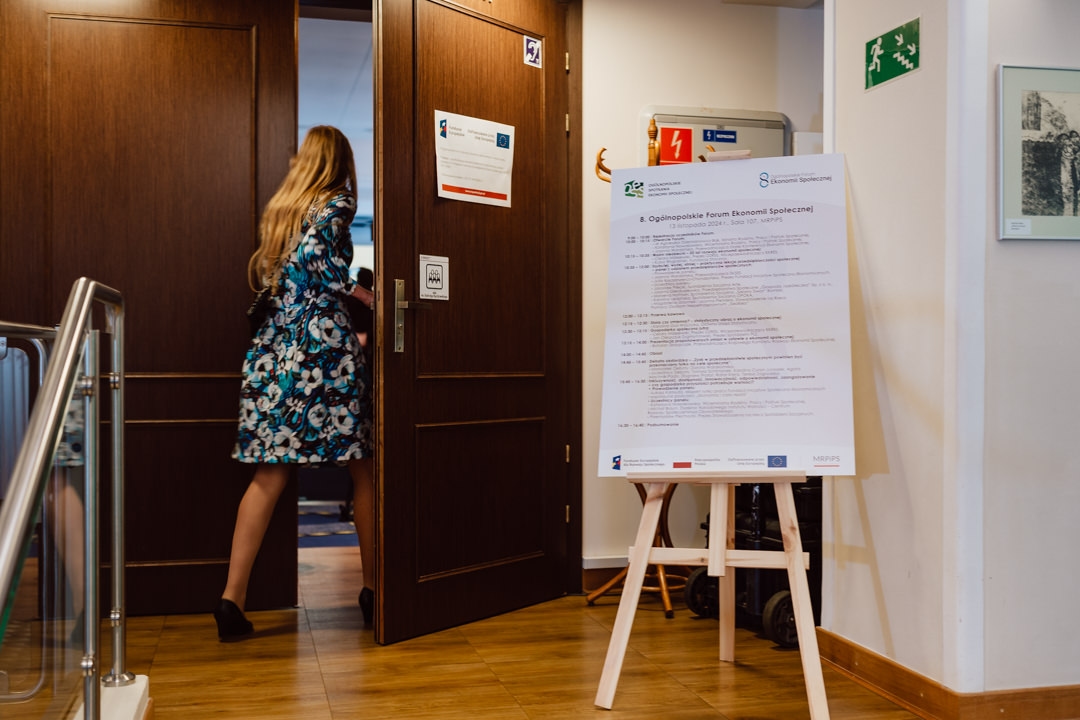 Eine Frau in einem geblümten Kleid betritt den Raum durch eine Holztür. Neben der Tür steht ein großer weißer Veranstaltungskalender auf einer Staffelei. Die Szene sieht aus wie ein Konferenz- oder Seminarraum.  