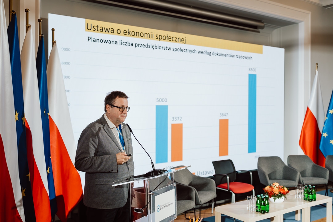 Ein Mann im Anzug spricht auf einem Podium vor einem Bildschirm, auf dem ein Balkendiagramm mit polnischem Text zu sehen ist. Mehrere polnische und EU-Flaggen sind auf beiden Seiten der Bühne aufgestellt und leere Stühle stehen in der Nähe. 