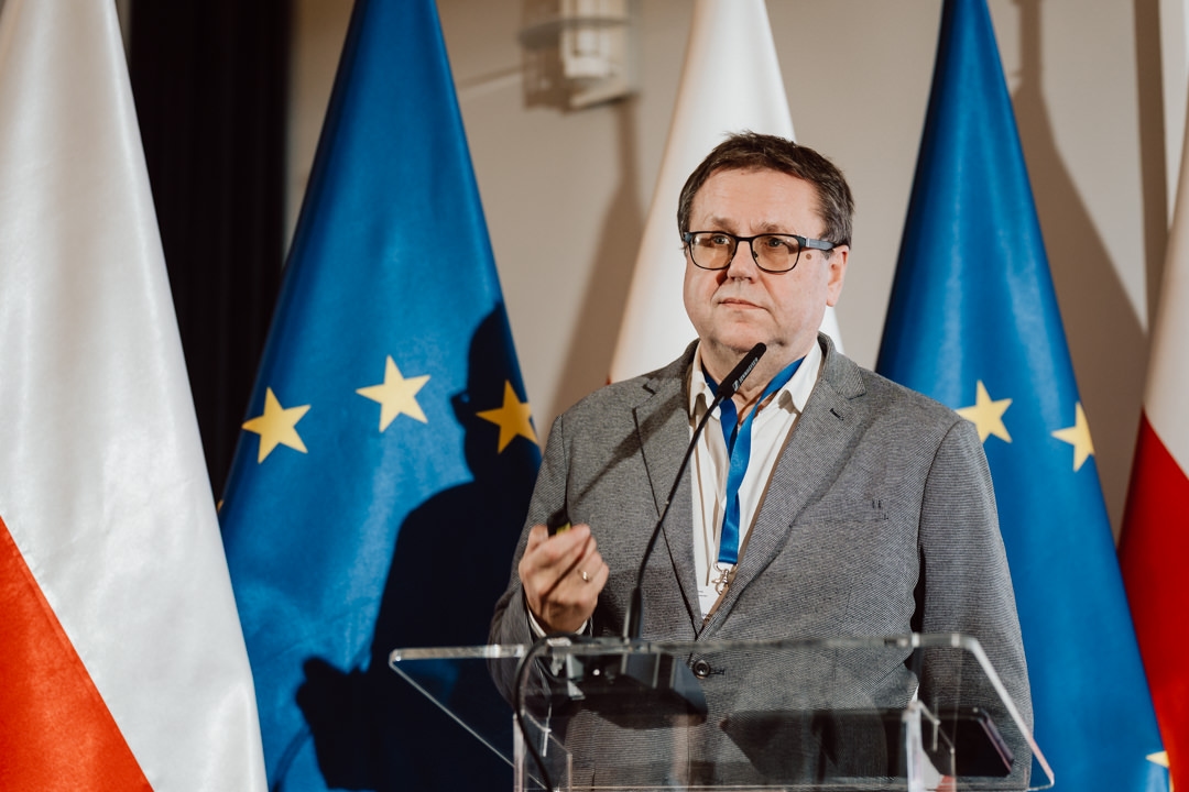 Ein Mann in einem grauen Anzug spricht an einem Podium mit Mikrofon vor den Flaggen Polens und der Europäischen Union.