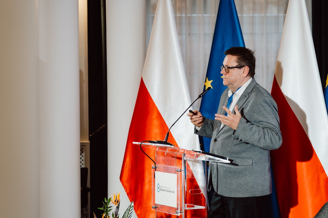 Ein Mann in einer grauen Jacke spricht auf einem transparenten Podium mit einem Mikrofon. Im Hintergrund sind die Flaggen Polens und der Europäischen Union zu sehen.