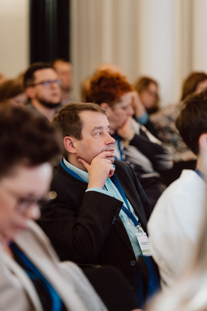 Ein Mann im Anzug mit einem blauen Schlüsselband sitzt inmitten einer Gruppe von Menschen und hört mit seiner Hand am Kinn aufmerksam zu. Es scheint sich um eine Konferenz oder ein Seminar zu handeln.