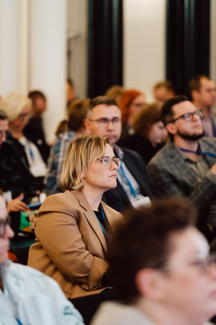 Eine Gruppe von Menschen sitzt dicht beieinander in einem Konferenzraum und hört aufmerksam einer Präsentation zu. Eine Frau in einer braunen Jacke und mit Brille steht im Mittelpunkt der Aufmerksamkeit der Zuhörer. 