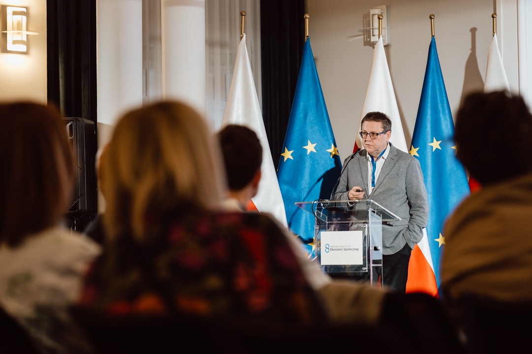 Ein Mann spricht auf einem hellen Podium vor den Flaggen der Europäischen Union und Polens und wendet sich an das im Vordergrund sitzende Publikum.