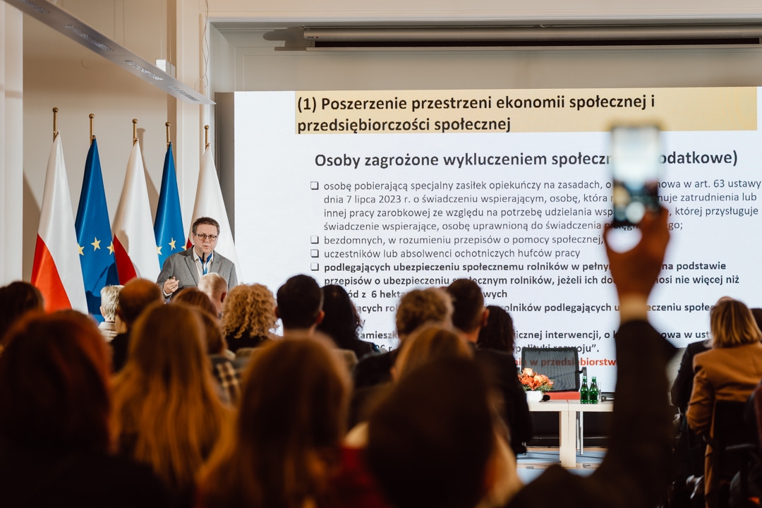 Der Redner wendet sich an die Zuhörer im Konferenzraum. Er steht in der Nähe von Fahnen und einem großen Bildschirm, auf dem Informationen in polnischer Sprache über die Sozialwirtschaft und Ausgrenzung angezeigt werden. Die Teilnehmer sitzen, einige heben ihre Hände oder machen Fotos. 