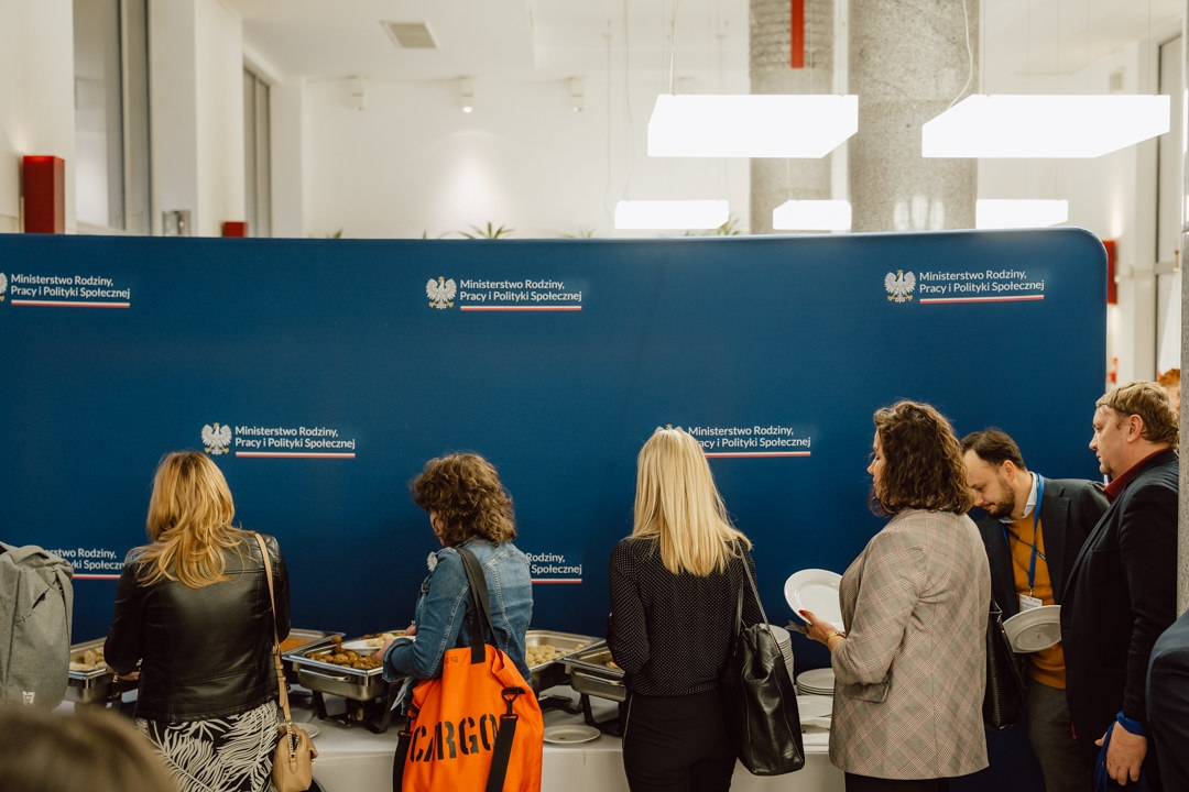 Eine Gruppe von Menschen steht vor einem blauen Hintergrund mit dem Logo des polnischen Ministeriums an einem Buffettisch an, hält Teller in der Hand und serviert sich das Essen. Einige haben Taschen oder Jacken dabei. Die Szene befindet sich in einem Raum mit heller Beleuchtung. 