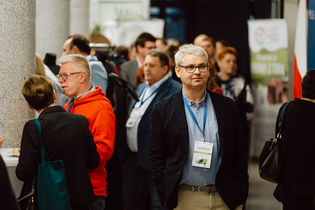 Eine Gruppe von Personen auf einer Konferenz oder einem Networking-Event, einige tragen Namensschilder und Schlüsselbänder. Ein Mann mit Brille steht im Vordergrund, während sich andere im Hintergrund unterhalten. 
