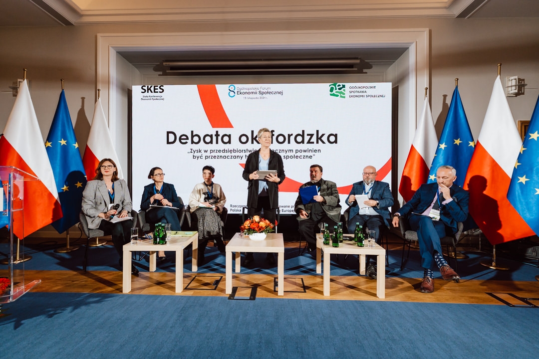 Sechs Personen sitzen auf der Bühne während einer formellen Podiumsdiskussion vor einem Bildschirm mit der Aufschrift 'Oxford Debate'. Hinter ihnen sind die Flaggen von Polen und der Europäischen Union zu sehen. Eine Frau steht in der Mitte und spricht.  