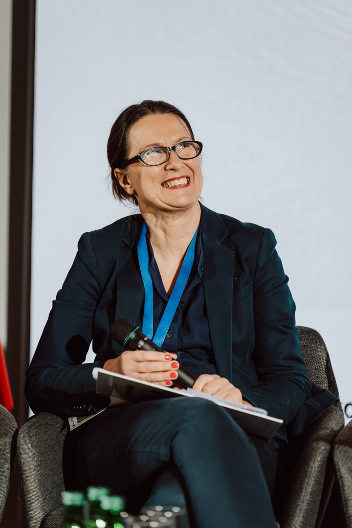 Eine Frau in einem marineblauen Anzug sitzt auf einem Stuhl und lächelt, sie hält ein Mikrofon und einen Notizblock in der Hand. Sie trägt eine Brille, hat roten Nagellack und ein Konferenzband um ihren Hals. Im Vordergrund sind Flaschen zu sehen.  