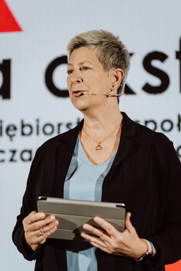 Eine Person mit kurzen Haaren, die eine Jacke und ein hellblaues Oberteil trägt, spricht auf der Bühne über ein Mikrofon in einem Headset und hält ein Tablet. Ein Teil des Textes und ein rotes Dreieck sind vor einem unscharfen Hintergrund zu sehen. 